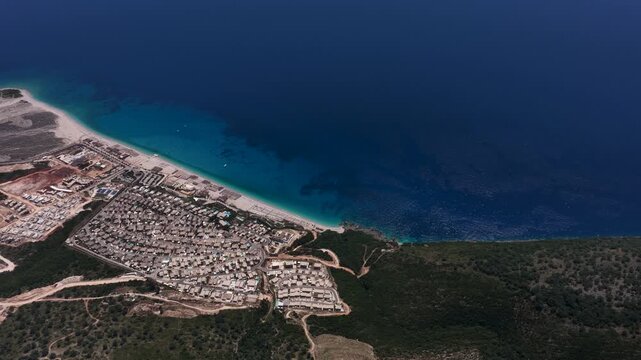 Aerial view of the turquoise waters and sandy beach of the Albanian Riviera with a resort village nestled at the foot of green mountains in Llogara, Vlore County, Albania.