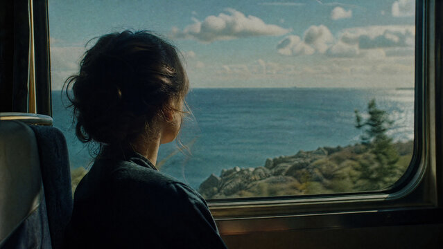 Back view of a woman looking out a train window at a rocky coastline. Female passenger enjoying the ocean view during a daytime journey. Travel and tourism concept