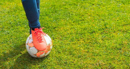 Female football player in bright pink cleats stands on a soccer ball on a green grass field, showcasing athletic footwear and sports equipment in a natural setting © Irina Flamingo