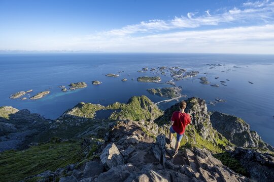 Mountaineer on the summit of Festvagstinden, view of the sea and archipelago islands with the village of Henningsv&aelig;r, coastal landscape, Lofoten, Nordland, Norway