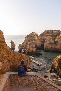 Female hiker sits on stone wall and looks over rocks in the sea, Algarve rocky coast, Ponta da Piedade, Lagos, Portugal