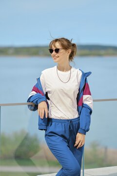 A woman stands confidently by the water, dressed in comfortable sportswear and sunglasses. She poses against a clear blue sky, enjoying the sunny day. The scene exudes a relaxed summer vibe