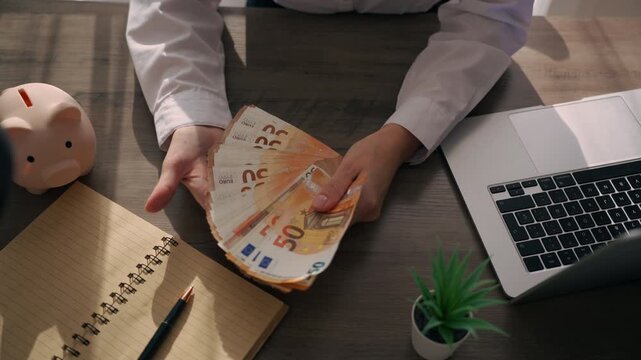 Businesswoman holding a stack of euro banknotes and gesturing with confidence at her office desk. Salary payment, income, financial reward and successful career concept.