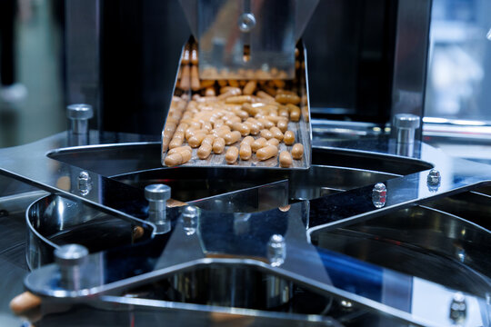 brown medicine capsules or dietary supplements falling from a metal chute into a rotary sorting machine. Pharmaceutical manufacturing and industrial mass production packaging process.