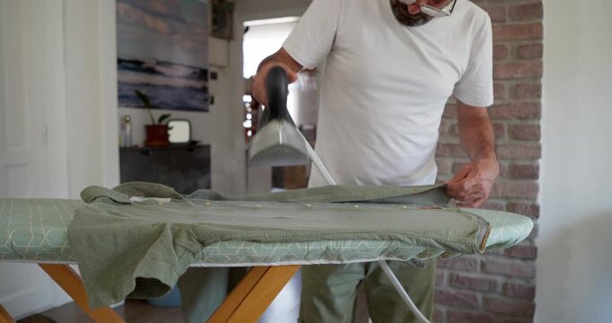 Man Ironing Shirt: A man in a white t-shirt and safety glasses is ironing a green shirt on an ironing board in a well-lit room, creating crisp creases for a neat appearance.