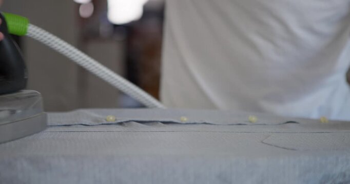 Steam ironing a shirt: Close-up shot of a person's hands carefully ironing a white shirt with a steam iron on an ironing board, demonstrating household chores and clothing care.