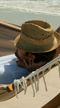 Vertical video: Reclining man in straw hat and white T propping head on beach hammock seeking rest