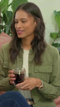 Vertical video: Laughing woman in olive shirt holding clear glass mug on pink sofa, toasting friend