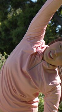 Vertical video: Lifting right arm, woman in pink top leaning left, holding side stretch to relax