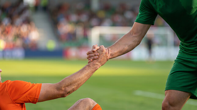 Close up shot of a soccer player in green uniform helping an opponent in orange jersey to stand up on a grass field after a match
