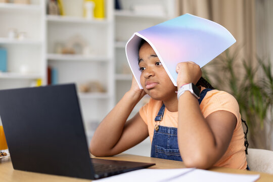 Black teenage girl sitting at desk and covering head with notebook while studying online with laptop. Concept of frustration, boredom, academic stress, and learning challenges