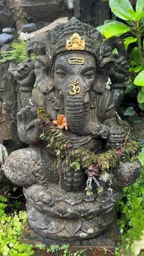 Vertical shot of a stone sculpture of Ganesha surrounded by decorative plants in an outdoor cultural setting. Sacred monument.