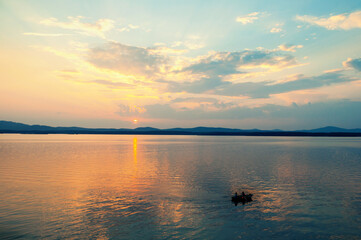 Obraz premium Summer vacation landscape, the sun setting behind the mountains and water surface lit by sunset and an unidentified people on a boat at the distance
