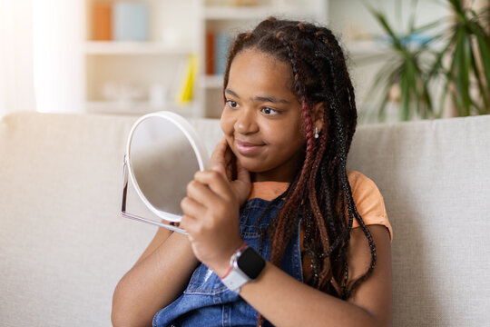 Black teenage girl looking at her reflection in handheld mirror at home. Smiling young female checks her appearance during a self care and personal grooming routine