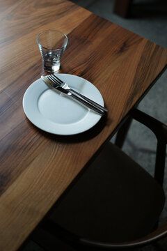 High angle shot of a clean white ceramic plate with cutlery and an empty glass on a natural wood dining table. Ready for serving in a cozy modern restaurant