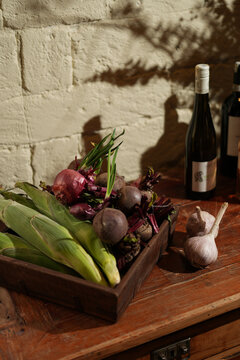 Rustic harvest scene with fresh corn, beetroots, red onions in a wooden box, garlic, and wine bottles on a table. White brick wall with tree shadows