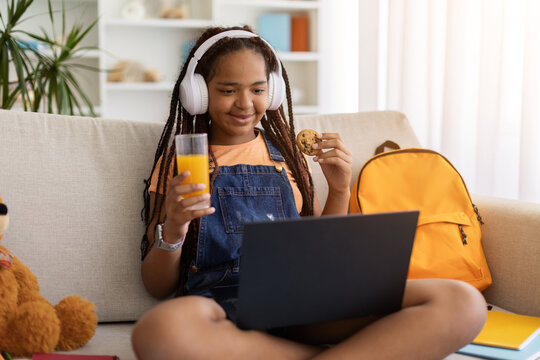 Black teenage girl enjoying cookie and orange juice while using laptop at home. Student taking relaxing break during online learning and study activities