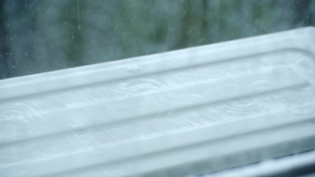 Window view of outdoor air conditioner unit with raindrops falling, splashing on metal surface. Rainy weather seen from inside. Window view of outdoor AC unit with raindrops falling
