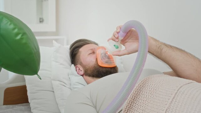 Pull out of man lying in hospital bed and holding oxygen mask over face while breathing. Ideal for medical training videos, healthcare ads, or anesthesia procedure concept