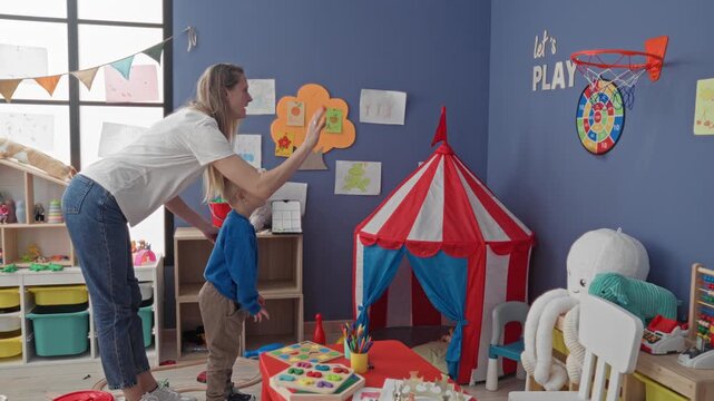 Woman stands watching boy reaching into a red and white striped play tent among scattered toys in a bright playroom inside a building; playful care.