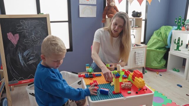Woman helps little boy stack toy blocks, hand reaches toward pieces on a play table in building; learning calm growth.