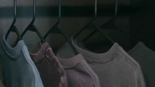 Man hand selecting outfit from row of colorful t shirts hanging on black plastic hangers inside dark closet, representing daily routine of getting dressed, choosing clothes. 
