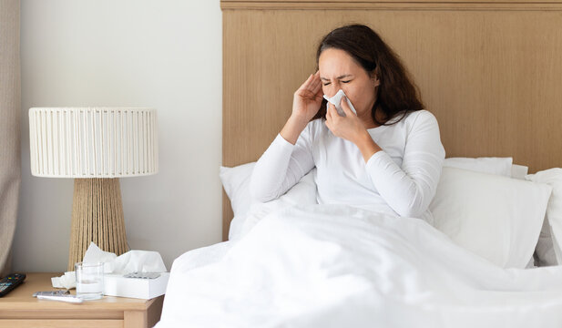 Sick woman sneezing into a tissue while sitting in bed, ill adult female showing symptoms of cold, flu or seasonal allergy during recovery at home