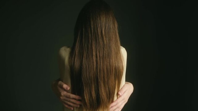 Woman with long brown hair, seen from behind, wrapping her arms around herself in self love hug, dark background, symbolizing comfort, care, mental health. Woman hugging herself from behind