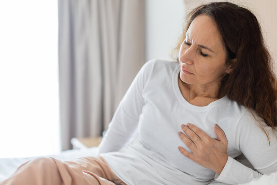 Woman sits on bed holding hand to chest with pained expression, chest pain or heartburn concept in bright bedroom for healthcare and cardiology lifestyle commercial use