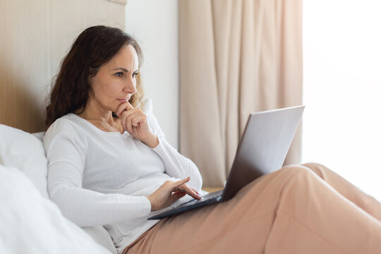 Thoughtful adult woman working on laptop while sitting in bed, focused on online tasks, planning or research in a comfortable bedroom environment