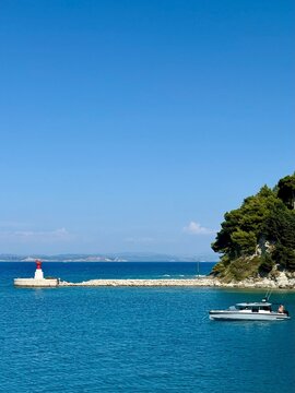 Vertical scenic view of a red and white lighthouse on a stone pier with a modern motorboat yacht floating on turquoise sea water at Sazan Island coast, Albania.