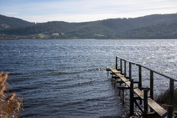 Fototapeta premium Weathered wooden jetty over a windy, sunlit river against a backdrop of green Tasmanian hills