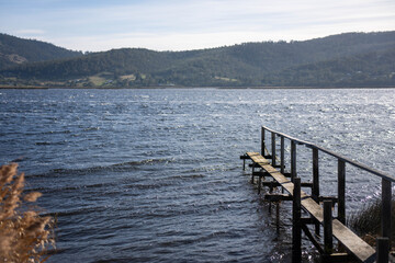 Fototapeta premium Weathered wooden jetty over a windy, sunlit river against a backdrop of green Tasmanian hills
