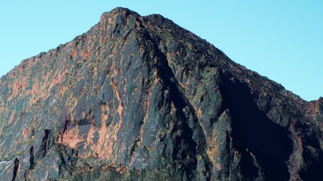 A large mountain rises sharply against a clear sky. The surface shows many colors and textures. The sunlight highlights its features well during mid day.