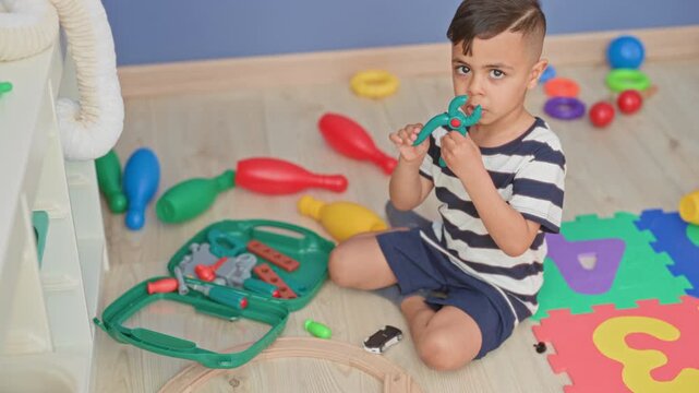 Little boy playing with green toolkit and toy car, holding pieces on foam alphabet mat inside building; concentration.