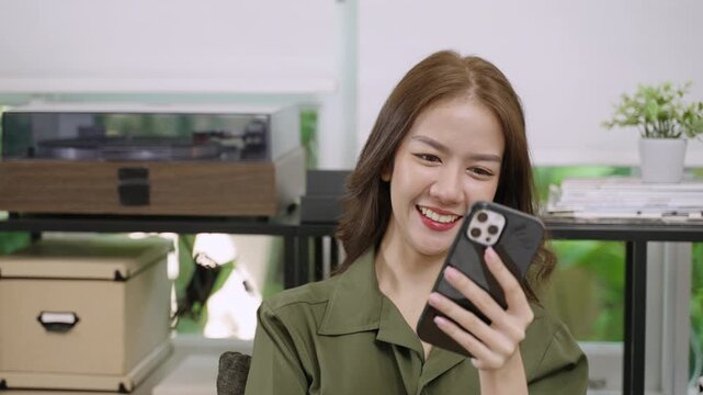 Happy attractive young Asian woman is using smartphone app for chatting and video calls online while resting and relaxing on break time from working at her desk in office. Female freelancer with cell.