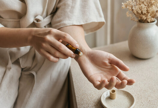 Person applying essential oil from a small amber glass roller bottle onto their wrist, with a vase of dried flowers on a beige stone surface