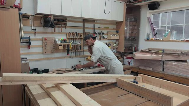Focused young carpenter preparing for manual labor putting yellow pencil behind his ear while grabbing tape measure and try square at workbench in bright rustic workshop authentic wide shot