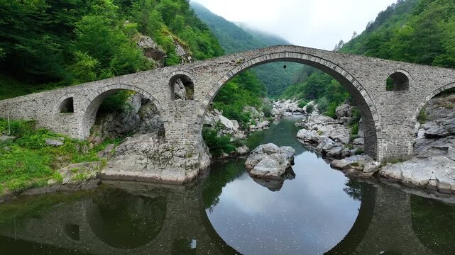Devil's Bridge in Ardino, Rhodope Mountains, Bulgaria. Low drone flight over the Arda River, passing under the main arch of an ancient stone bridge representative of Ottoman architecture. Europa above