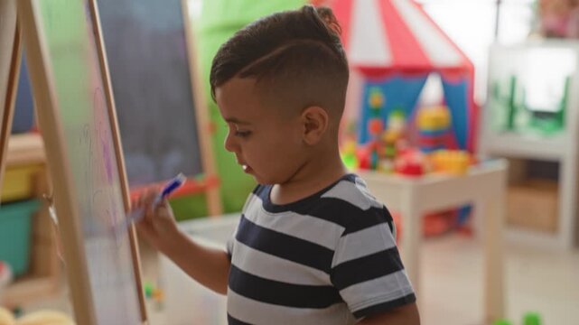 Little boy drawing on whiteboard at easel, hand holding blue marker, wearing striped shirt, surrounded by toys in studio; curiosity learning.