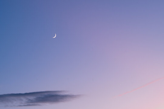 Slender crescent moon in a pastel twilight sky with a single dark cloud.