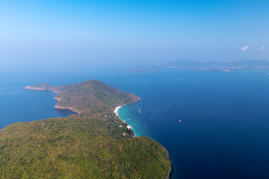 Tropical Ko He Coral Island off Phuket, Thailand, features lush green jungle, white sandy beaches, and clear blue ocean with anchored boats under a bright sky