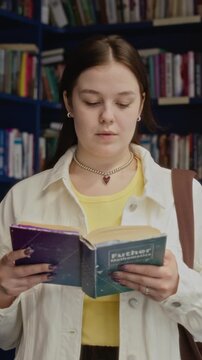 Vertical medium shot of Caucasian teenage girl standing in library, reading mathematics textbook. Clip ideal for promoting high school education, academic tutoring