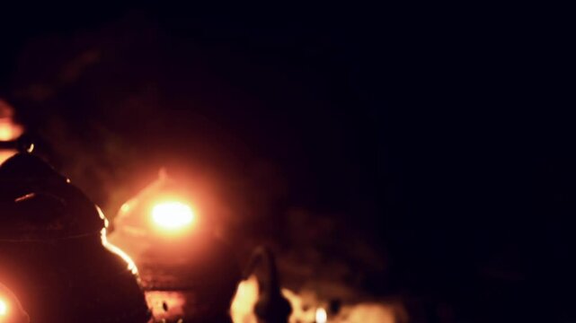 Candles are lit and placed along a path in a dark area. The flickering light creates a warm glow while night surrounds. People gather around for a serene experience.