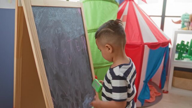 Little boy erases chalk scribbles on a wooden easel chalkboard while standing beside a red play tent in a bright playroom; creative concentration.
