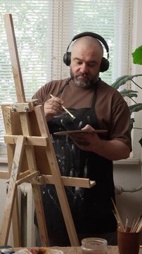 Portrait of a Caucasian, bearded millennial man wearing headphones, standing near an easel, wearing an apron, holding a palette, looking at a painting, and dancing. Concept: Hobbies and leisure