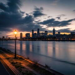 Fototapeta premium Illuminated streetlamp casts a warm glow along a riverside promenade facing a metropolitan skyline at twilight