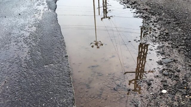 Reflections of electric poles, power lines, and trees shimmering in rainwater collected in a roadside pothole, creating an abstract urban landscape after rainfall

