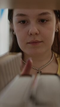 Vertical shot of Caucasian teenager browsing books on library shelf, selecting one and walking away. Clip ideal for high school or university enrollment campaigns