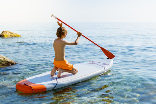 Young boy in orange shorts is kneeling on an inflatable stand up paddleboard in the shimmering clear sea. Holding a paddle